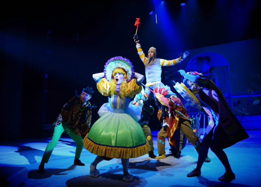 A young actress stands on stage in a colourful dress and hat, surrounded by her castmates during a musical number.