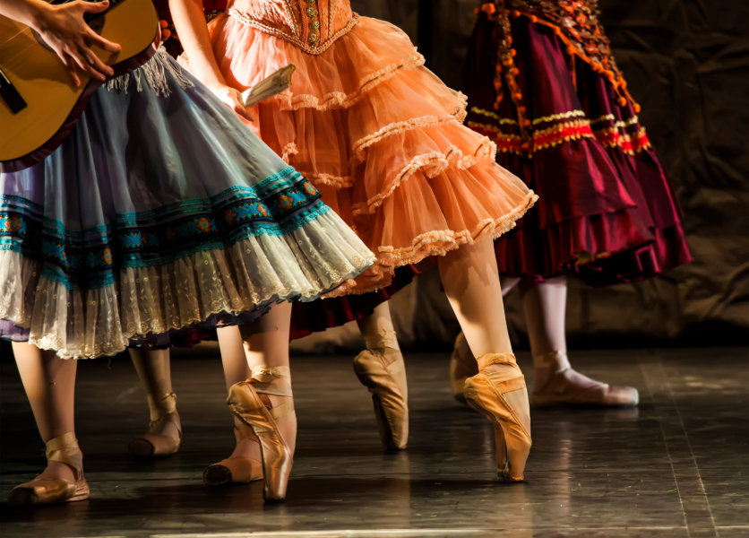 Performers wearing ballet shoes and ruffled dresses dance on stage while one plays music on a guitar.