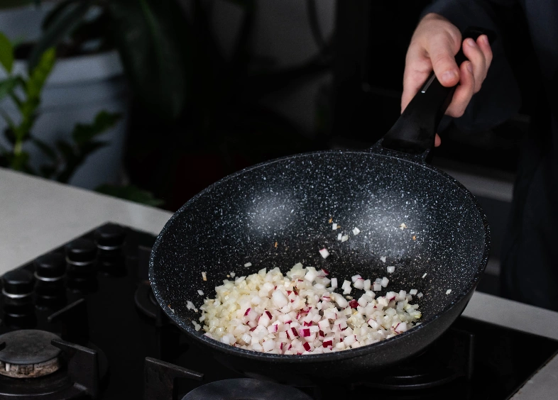 Close-up of a person cooking roasted onion and garlic in a frying wok pan on a gas stove.