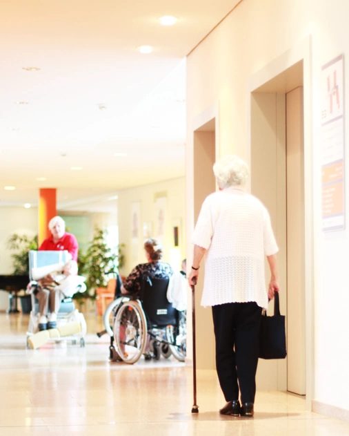 Old lady with walking stick in white shirt and black trousers walking past elevators towards reception area of a healthcare facility.